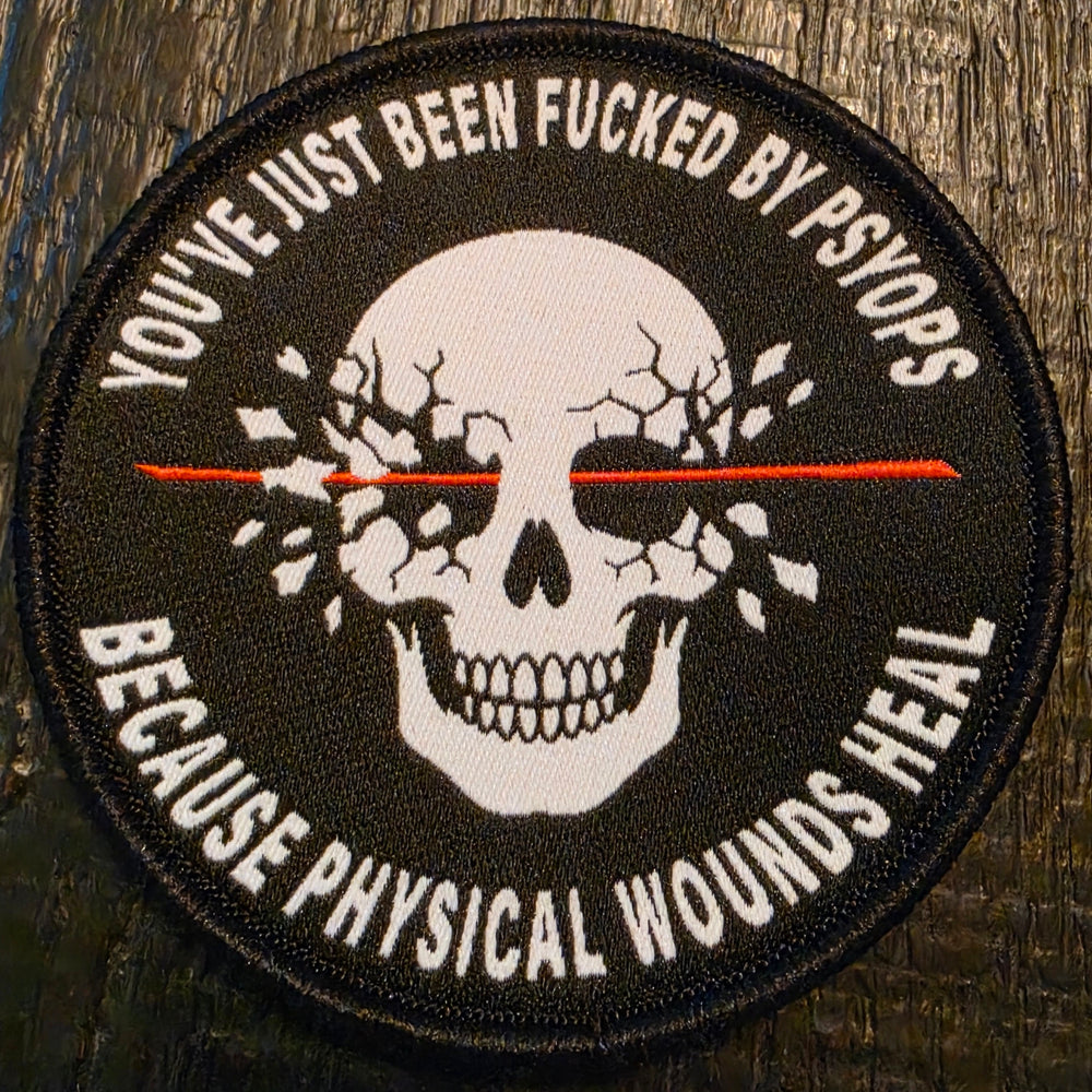 A circular embroidered patch featuring a white skull with cracked eyes and a red horizontal line through the center, surrounded by black text reading 'YOU'VE JUST BEEN FUCKED BY PSYOPS BECAUSE PHYSICAL WOUNDS HEAL' on a dark wood background.