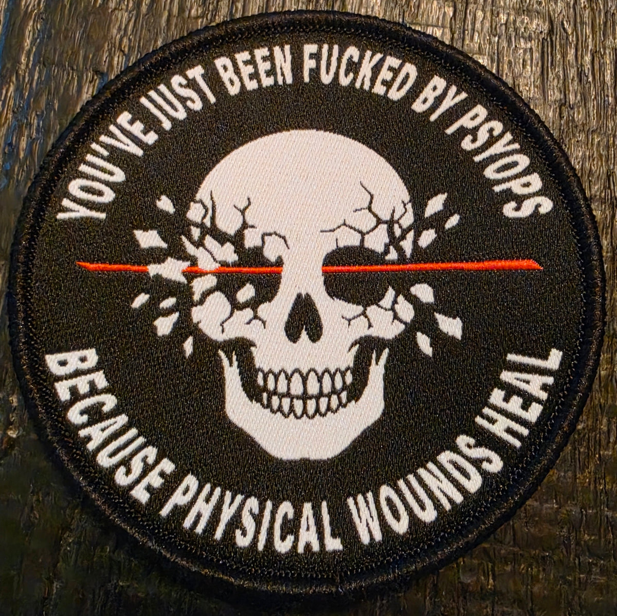 A circular embroidered patch featuring a white skull with cracked eyes and a red horizontal line through the center, surrounded by black text reading 'YOU'VE JUST BEEN FUCKED BY PSYOPS BECAUSE PHYSICAL WOUNDS HEAL' on a dark wood background.